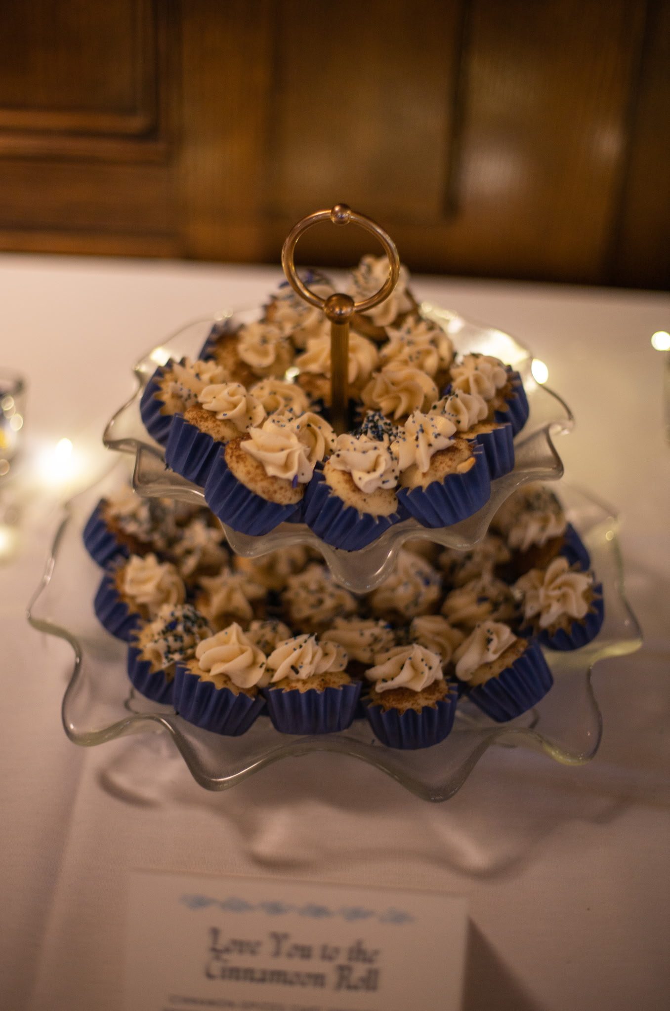 Two-tier stand of cinnamon roll cupcakes in navy wrappers with a gold ring topper